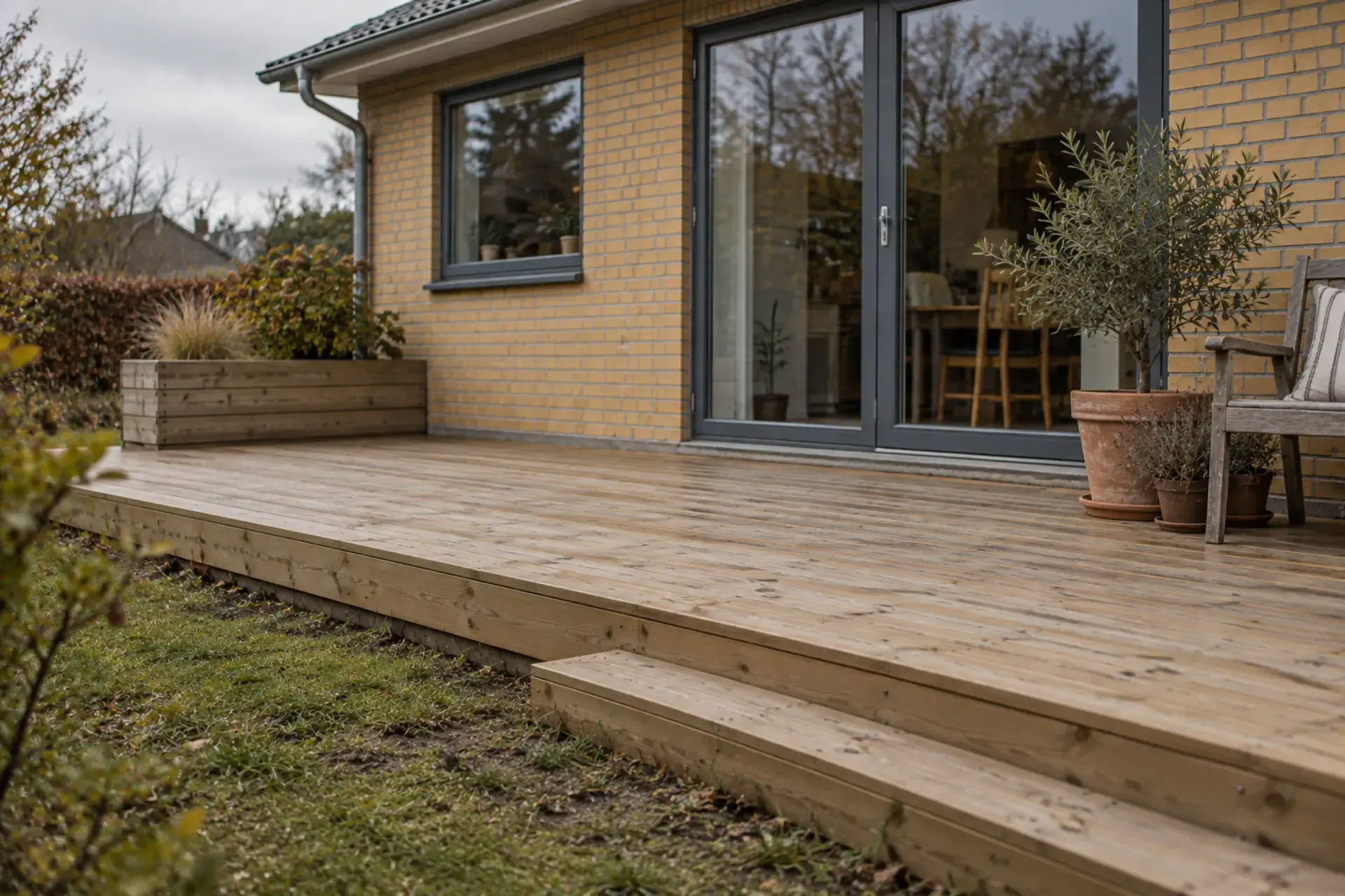 Træterrasse foran hus med store glasdøre og planter.